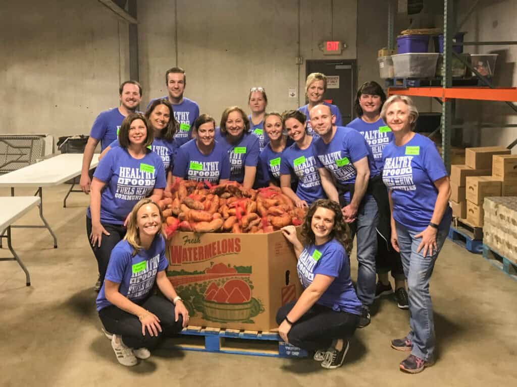 A group of people wearing matching blue Healthy Food T-shirts pose and smile around a large box filled with sweet potatoes in a warehouse setting.