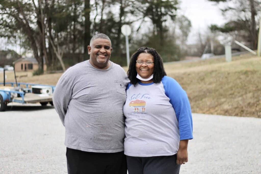 A smiling man and woman stand side by side outdoors on a cloudy day, both wearing casual shirts. Trees, grass, and a trailer are visible in the background.
