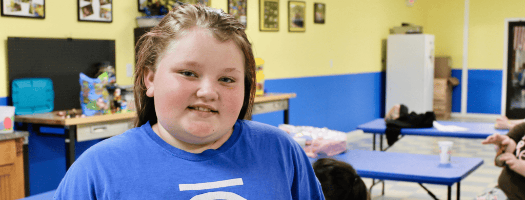 A young person with light skin and brown hair, wearing a blue shirt, smiles at the camera in a brightly colored room with yellow and blue walls, tables, and various items in the background.