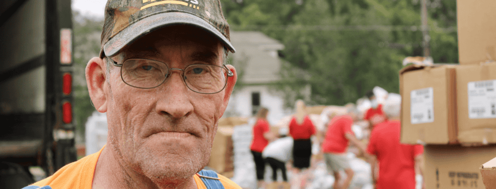 An older man wearing glasses, a cap, and overalls stands in front of boxes and a group of people working outdoors, possibly at a donation or distribution event. The background is slightly blurred.
