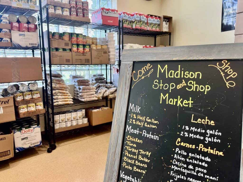 A chalkboard sign lists grocery items in English and Spanish inside a small market. Behind it are shelves stocked with canned goods, pasta, and boxes. Sunlight shines through a window onto the organized shelves.