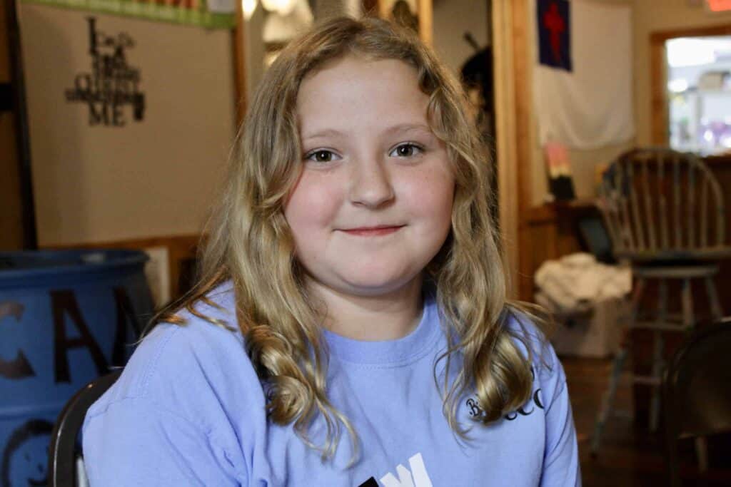A young girl with long, wavy blonde hair and a light purple shirt smiles while sitting indoors. The background shows chairs, a table, a blue barrel, and wall decorations.