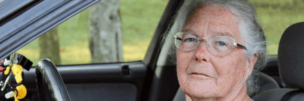 An older woman with gray hair and glasses sits in the drivers seat of a car, looking at the camera. The background shows trees and grass outside the car window.