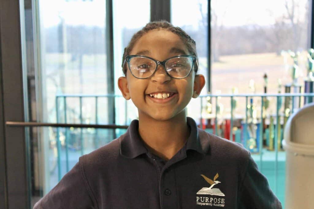 A smiling child with glasses wearing a dark blue Purpose Preparatory Academy shirt stands indoors with trophies and large windows in the background.