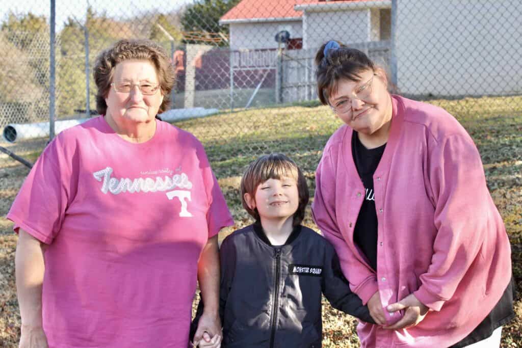 Two older women and a young boy stand outdoors, smiling and holding hands. The women wear pink clothing, and the boy is dressed in a black jacket. A fence and buildings are visible in the background.