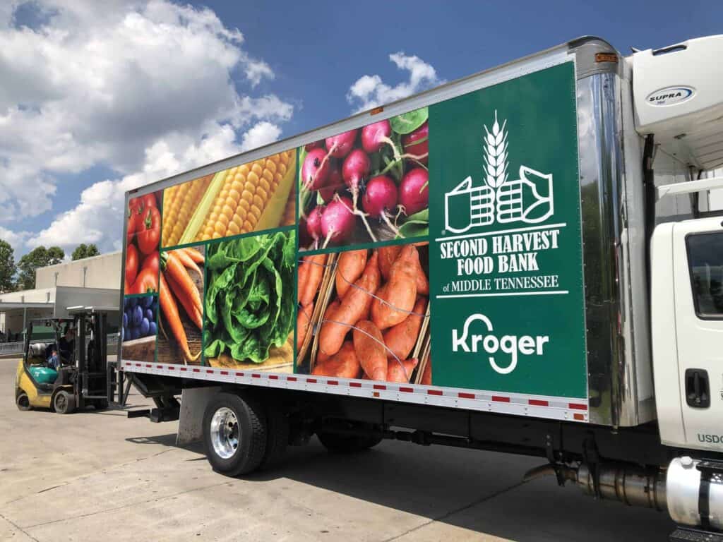 A truck displays colorful images of fresh vegetables and fruits, with the logos for Second Harvest Food Bank of Middle Tennessee and Kroger on its side. A forklift and building are visible in the background.