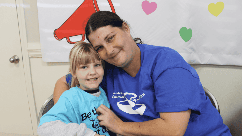 A woman in a blue shirt hugs and smiles with a young girl in a light blue shirt. They are seated in front of a white wall decorated with colorful hearts and a red megaphone cutout.