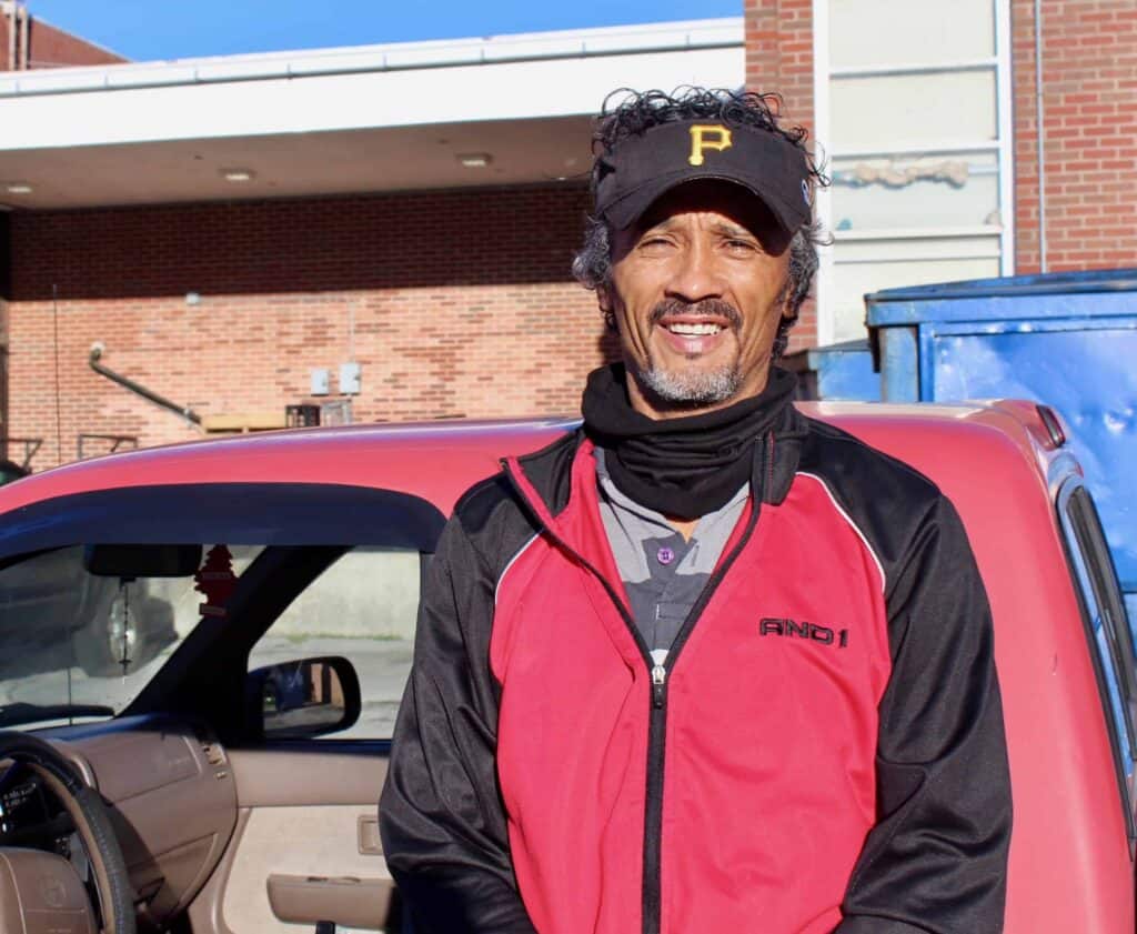A man wearing a black and red jacket and a black baseball cap with a yellow P stands smiling in front of a red vehicle, with a brick building and blue dumpster in the background.