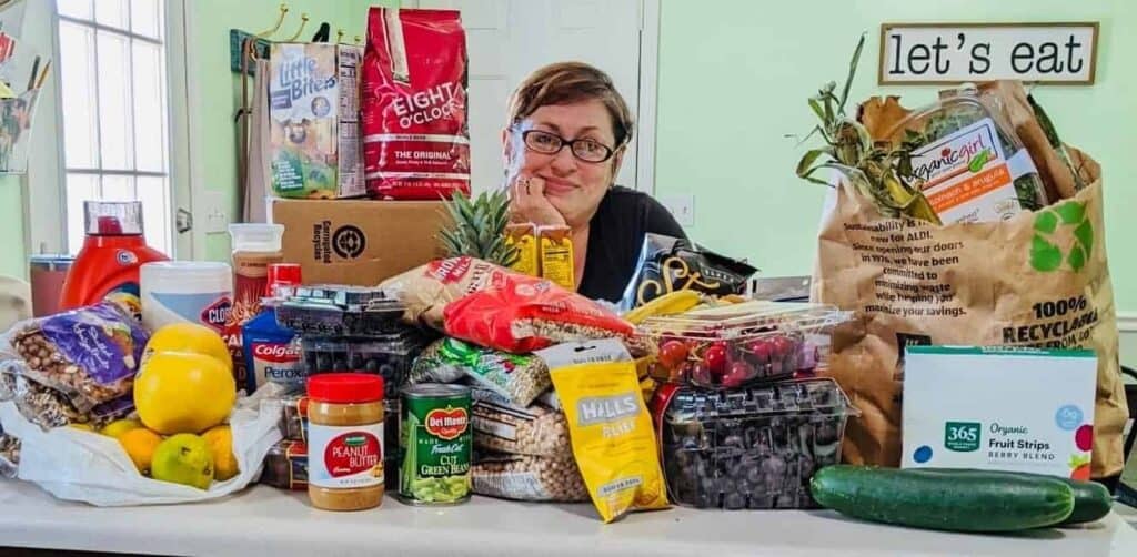 A person with glasses smiles behind a kitchen counter filled with various groceries, including fresh produce, canned goods, bread, snacks, and household items. A sign on the wall reads lets eat.