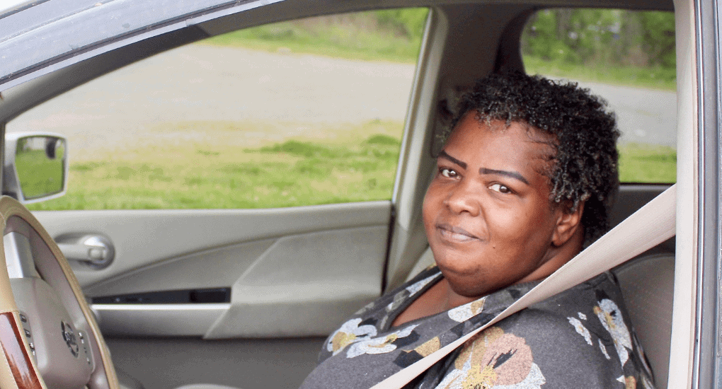 A woman with short curly hair sits in the drivers seat of a car, wearing a seatbelt and a floral-patterned shirt, looking at the camera and smiling slightly. Green grass and trees are visible outside the car window.