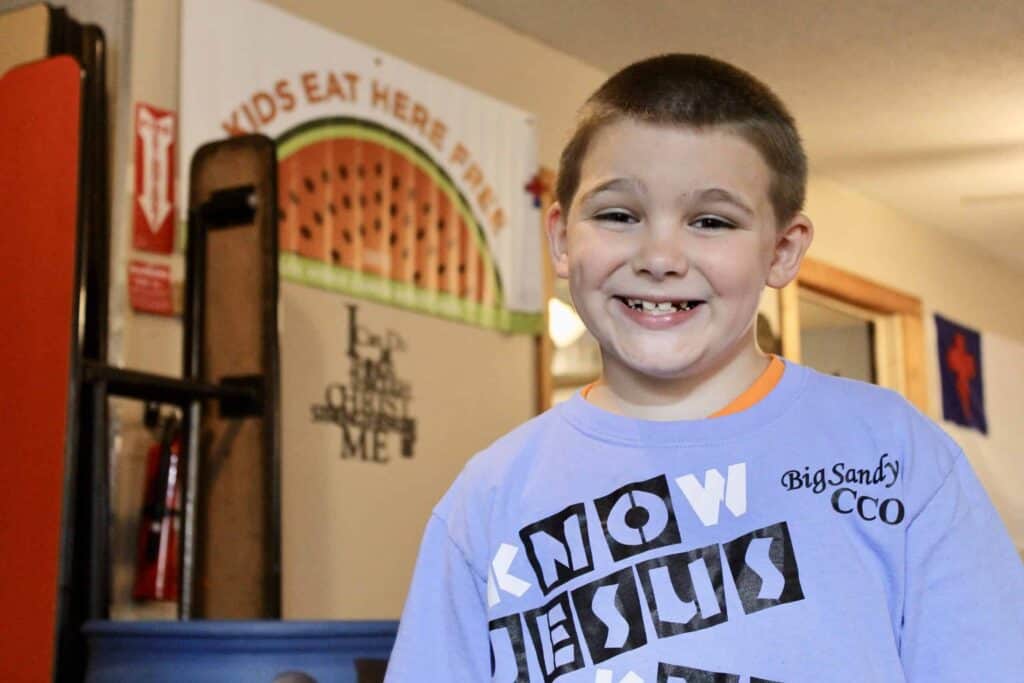 A young boy with short hair smiles at the camera, wearing a light blue shirt that says KNOW JESUS and Big Sandy CCO. A sign behind him reads Kids Eat Here Free above a watermelon illustration.