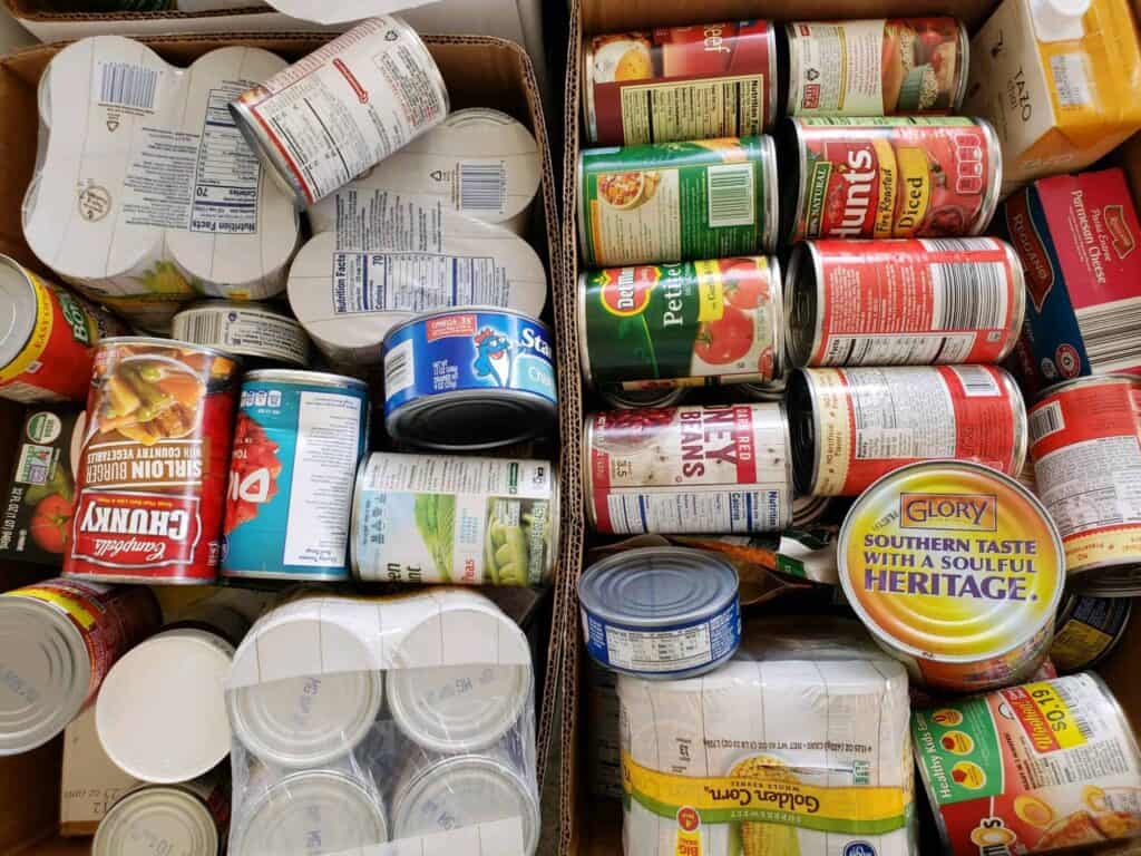 Two cardboard boxes filled with assorted canned foods, including vegetables, beans, soup, fruit, and tomato products, stacked closely together. Some cans are upside down or on their sides, showing colorful labels and tin tops.