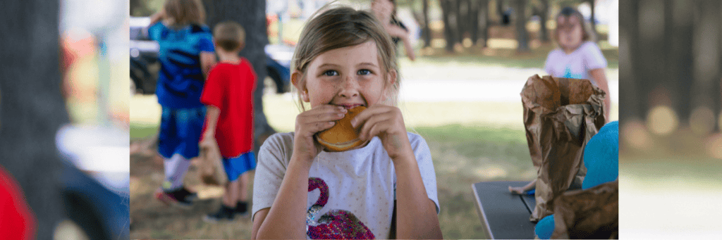 A young girl with light brown hair and a flamingo shirt smiles while eating a sandwich outdoors, with other children and lunch bags visible in the background.