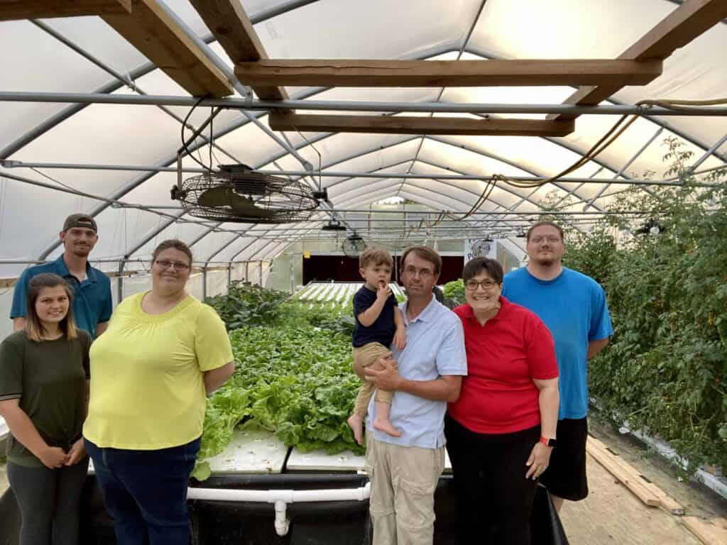A group of seven people, including a child, stand and smile inside a greenhouse filled with lush green plants and vegetables. The structure has a transparent roof and wooden beams overhead.