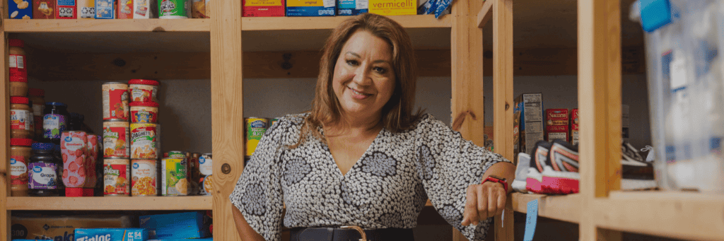 A woman stands smiling in front of wooden shelves stocked with canned goods, boxed food, and a pair of sneakers, possibly in a food pantry or storage room.