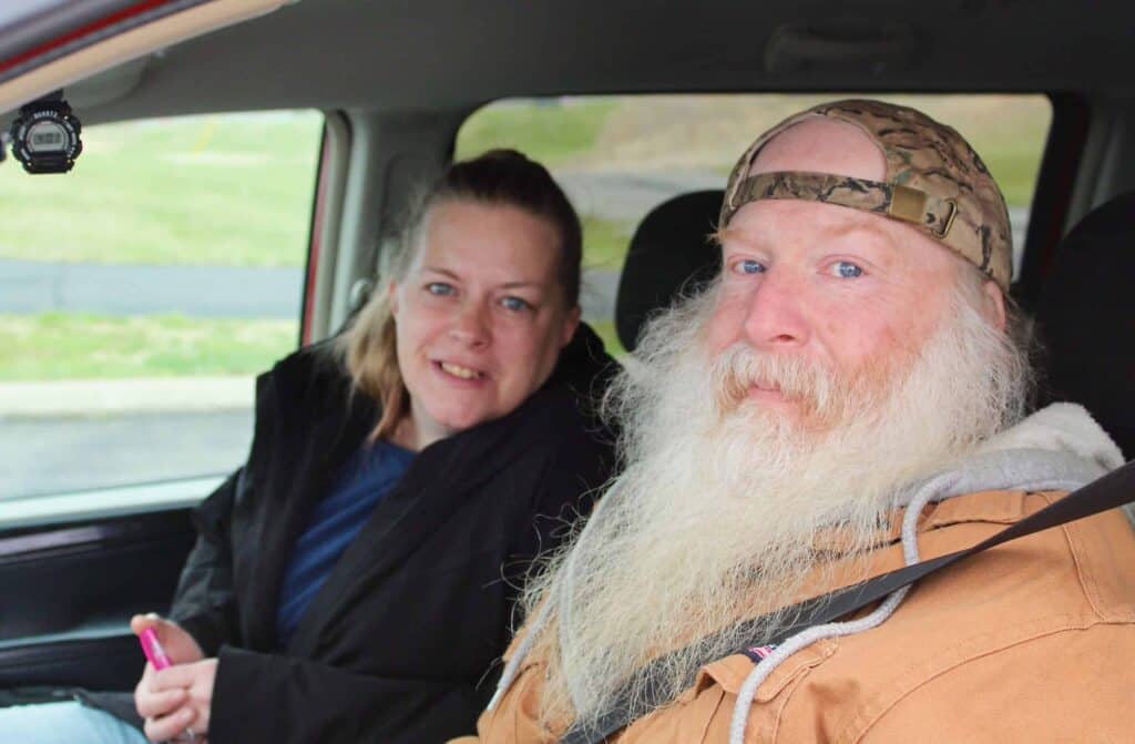 A woman and a bearded man wearing a camouflage cap and brown jacket sit together in the front seats of a car, both looking at the camera and smiling slightly.