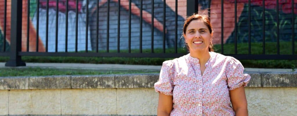 A woman wearing a light-colored floral blouse stands outdoors in front of a stone wall and a black metal fence with a colorful mural in the background. She is smiling and facing the camera.