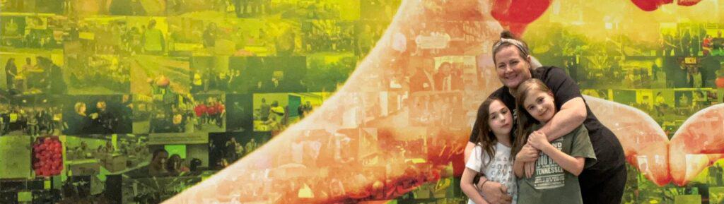 An adult and two children pose together, smiling, in front of a large, colorful collage mural featuring photos and abstract shapes in yellow and green tones.