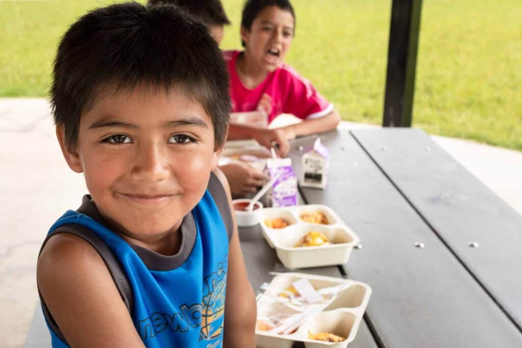 A young boy smiles at the camera while sitting at an outdoor picnic table with two other children, eating lunches from trays. The background shows green grass and a shaded area.