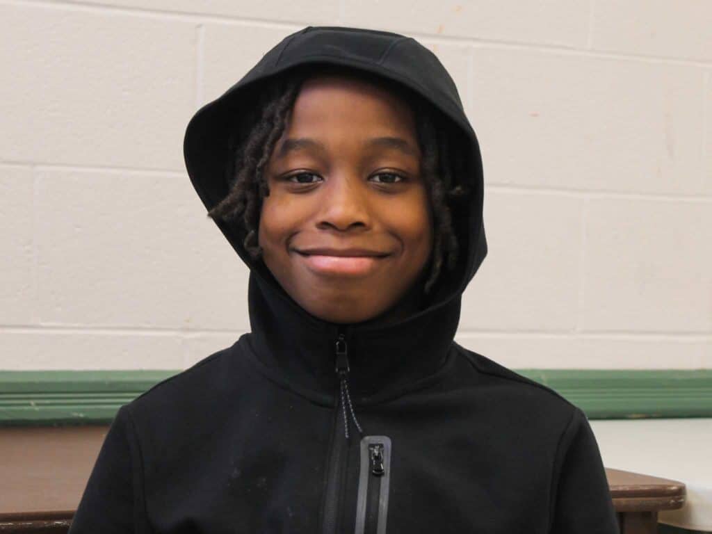 A young boy with medium-length braided hair smiles while wearing a black hoodie zipped up to his neck, standing in front of a white brick wall with a green trim.