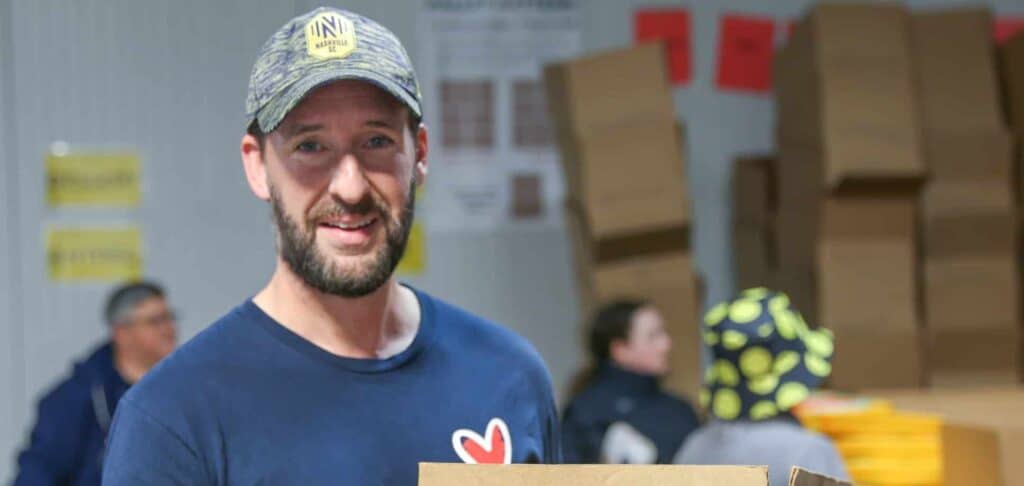 A smiling man in a cap and blue shirt with a red heart logo holds a cardboard box in a room with stacked boxes and people in the background.