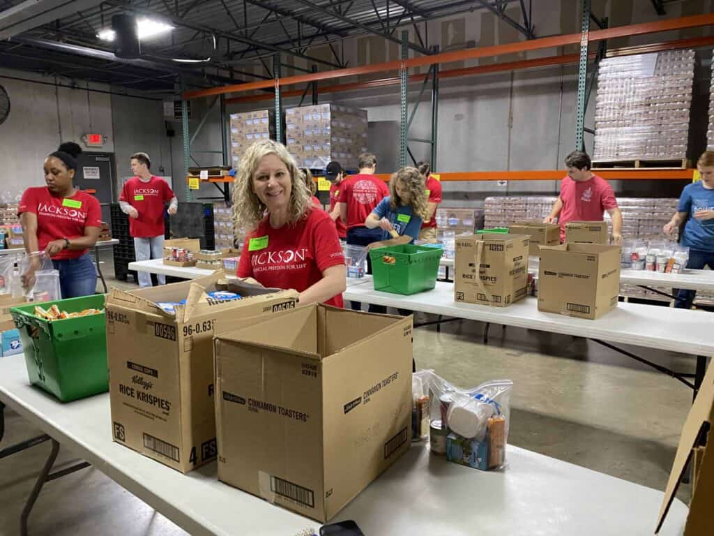 Volunteers wearing red shirts pack food and supplies into boxes at long tables in a warehouse. Shelves with boxes and bags are visible in the background. One woman smiles at the camera while working.
