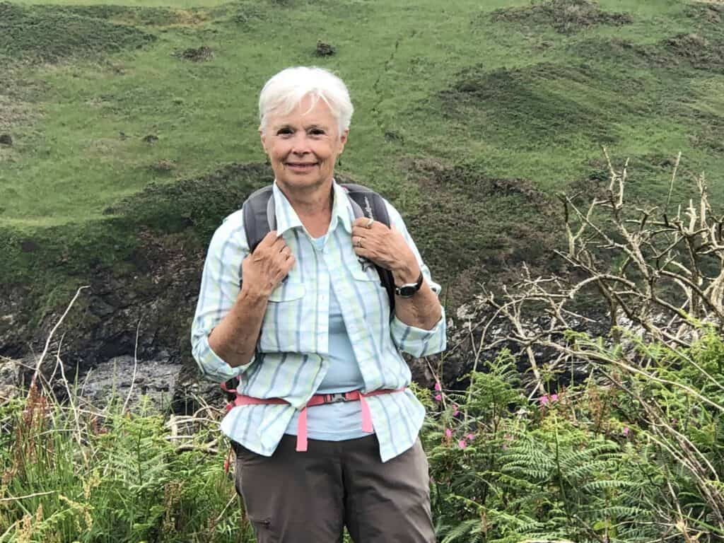 An older woman with short white hair wearing a plaid shirt, blue t-shirt, and backpack stands outdoors in a green, hilly landscape, smiling at the camera.