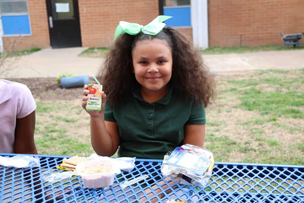 A young girl with curly hair and a large green bow sits at a blue outdoor table, smiling and holding up a juice box. In front of her is a school lunch with crackers, applesauce, and a packaged meal.