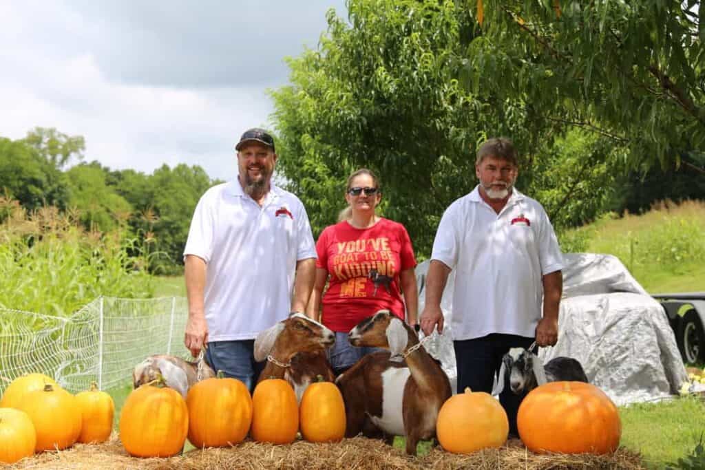 Three people stand outdoors behind a row of pumpkins and goats. Two men wear white shirts and a woman in sunglasses wears a red shirt. Green trees and fields are in the background.