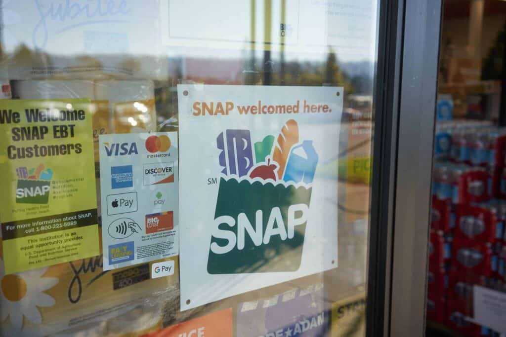 A glass door displays signs indicating that SNAP EBT, credit cards, and mobile payments like Apple Pay and Google Pay are accepted. A large sign reads “SNAP welcomed here” with a grocery bag graphic.