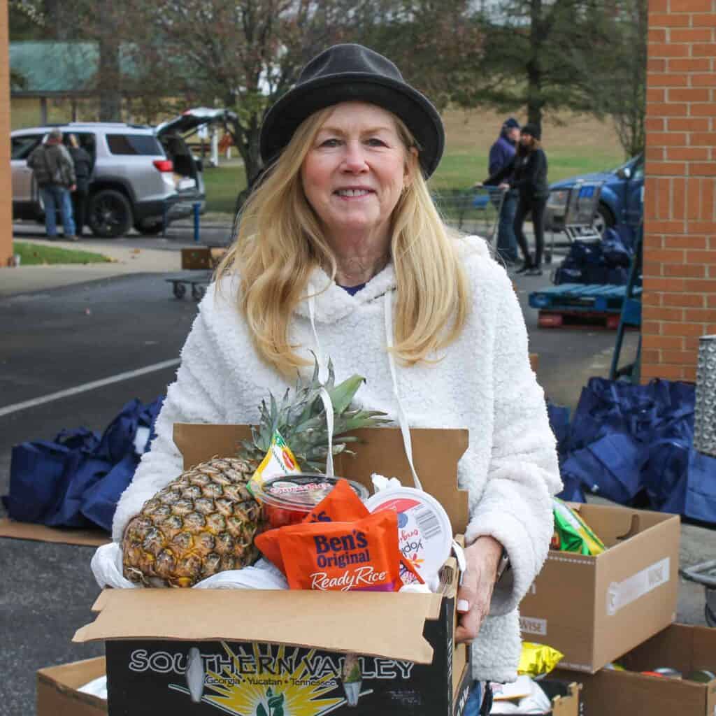 A smiling woman wearing a black hat and white jacket holds a box filled with groceries, including a pineapple, rice, and canned food, at an outdoor distribution event.
