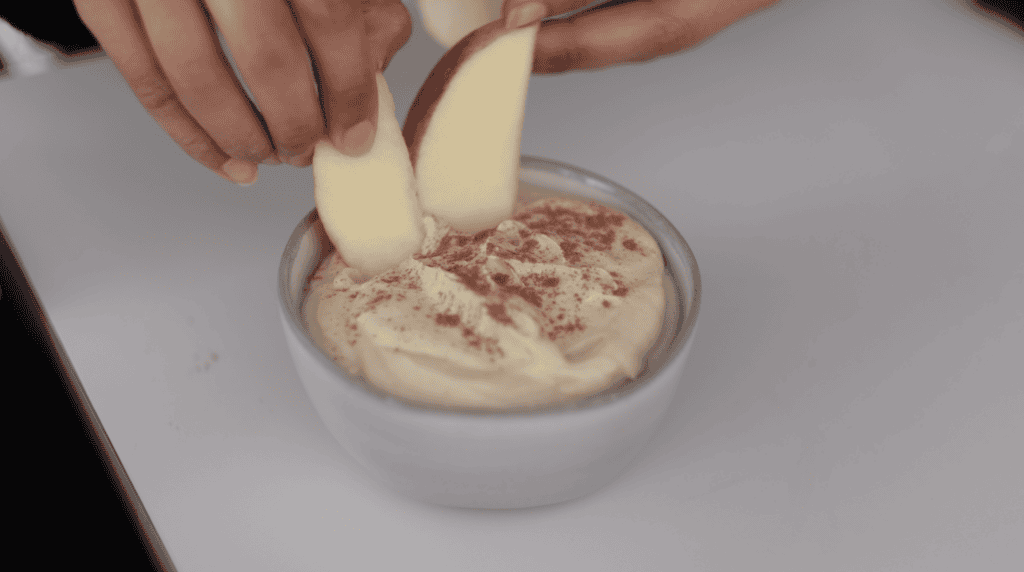 Two hands dip apple slices into a bowl of creamy dip topped with a sprinkle of cinnamon, set on a white surface.