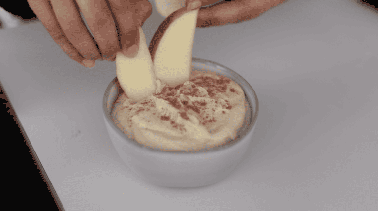 Two hands dip apple slices into a bowl of creamy dip topped with a sprinkle of cinnamon, set on a white surface.