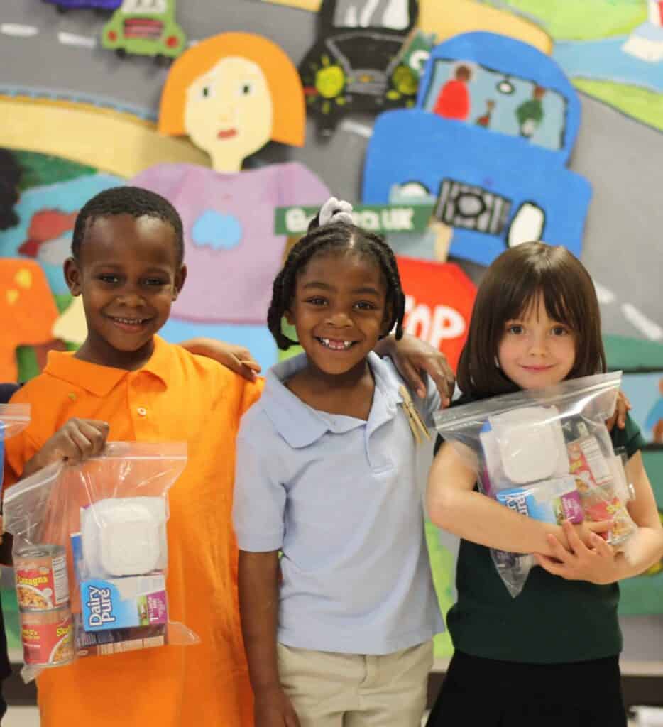 Three smiling children stand in front of a colorful mural, each holding a clear plastic bag containing food and drink items.
