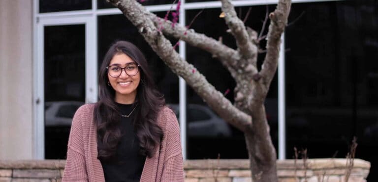 A woman with long dark hair and glasses, wearing a black shirt and mauve cardigan, smiles while standing outside near a leafless tree in front of a building with large windows.