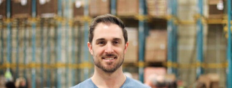 A man with short brown hair and a beard, wearing a blue shirt, stands smiling in front of tall warehouse shelves filled with stacked boxes.