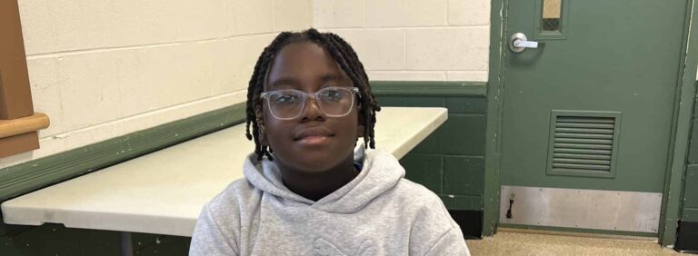 A child with glasses and short braids sits indoors at a table, wearing a light gray hoodie, with a green door and white-and-green walls in the background.
