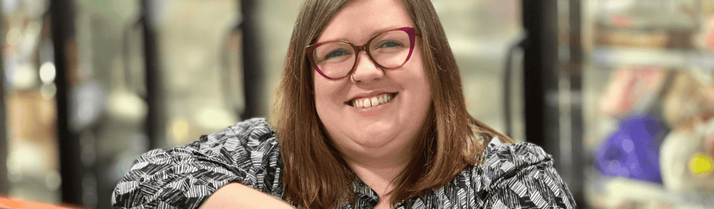 A smiling woman with brown hair and glasses sits indoors, wearing a patterned black and white shirt. The background is softly blurred, showing shelves with various items.