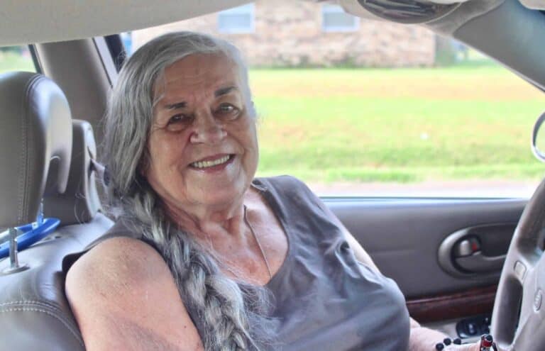 An older woman with long gray hair in a braid smiles while sitting in the driver’s seat of a car, wearing a sleeveless gray top. A grassy yard and brick building are seen outside the window.