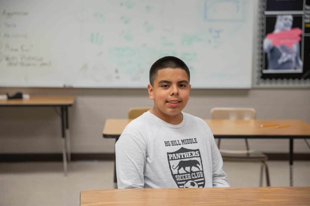 A boy with short dark hair wearing a Panthers Soccer Club shirt sits at a desk in a classroom with a whiteboard and bulletin board in the background.