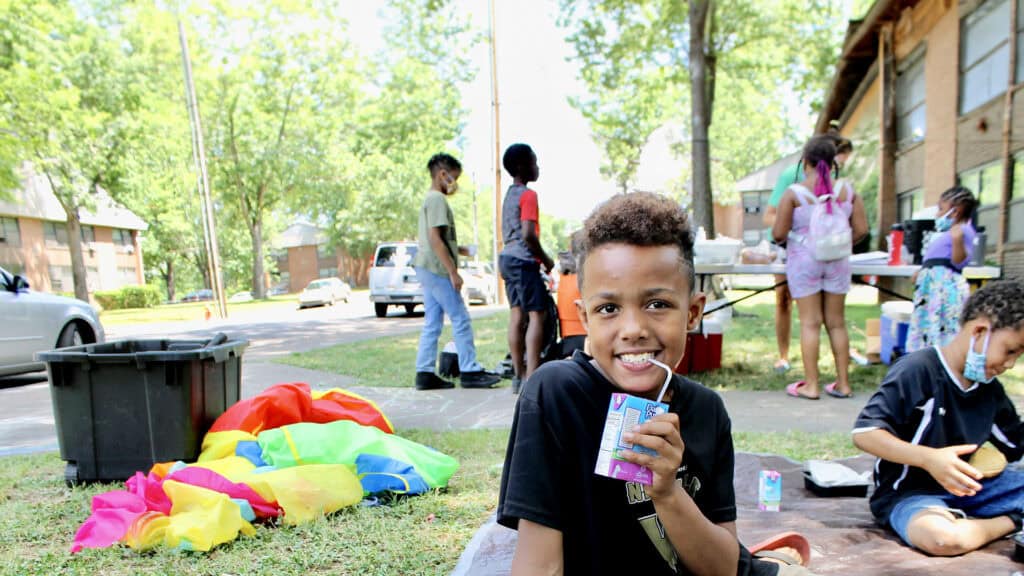 A smiling boy sits on the grass drinking juice, with colorful play items nearby. Behind him, children play and gather around tables outside on a sunny day, with trees and apartment buildings in the background.