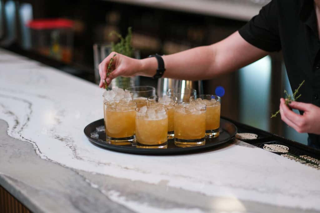 A person garnishes several cocktail glasses filled with ice and amber liquid on a black serving tray, placed on a marble bar counter.