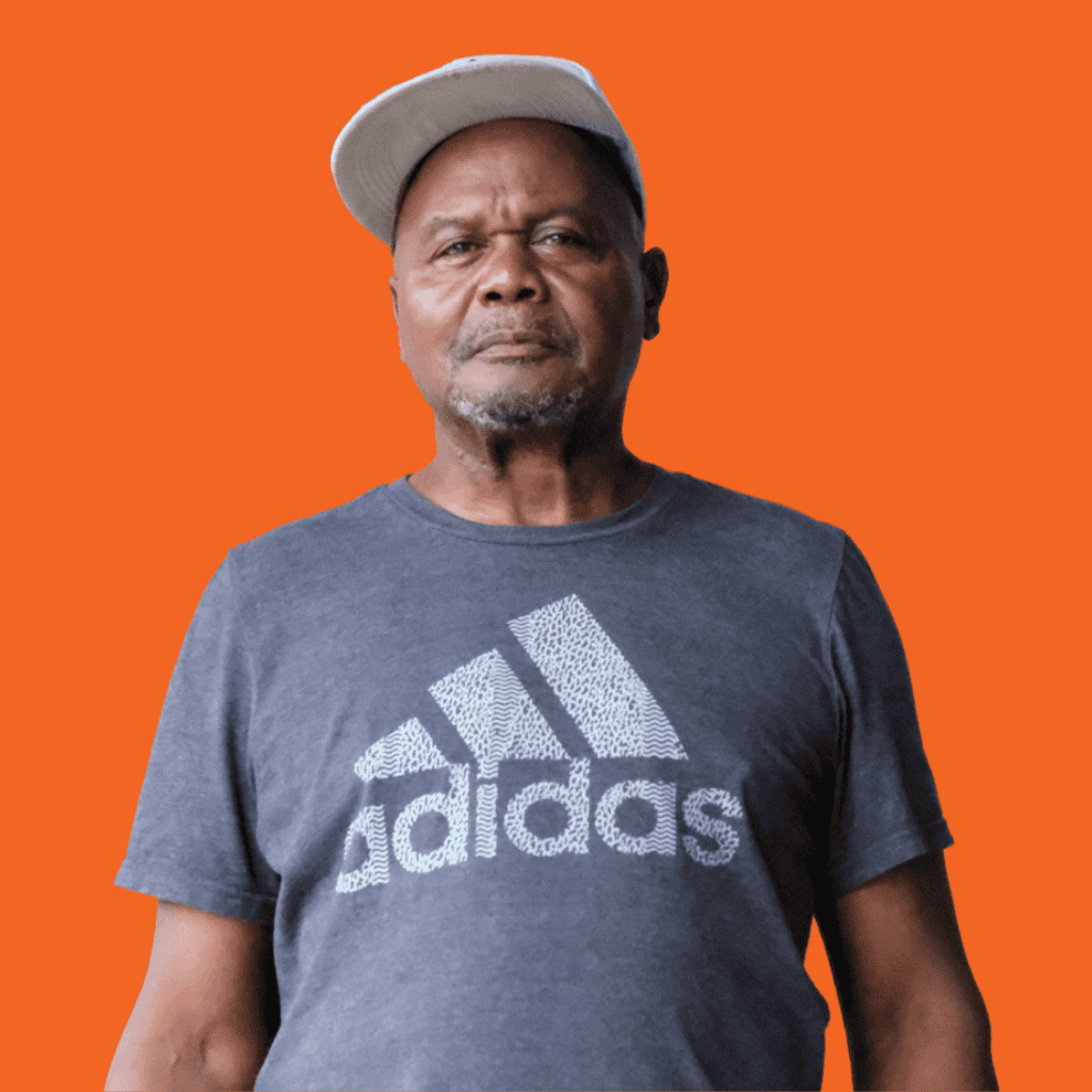 A man wearing a gray Adidas t-shirt and a light-colored cap stands in front of an orange background, looking straight ahead with a neutral expression.