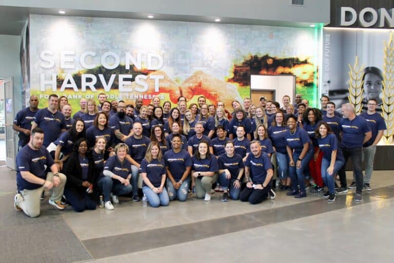 A large group of people wearing matching blue shirts pose and smile together inside the Second Harvest Food Bank of Middle Tennessee building. A mural and the words “Second Harvest” are visible on the wall behind them.