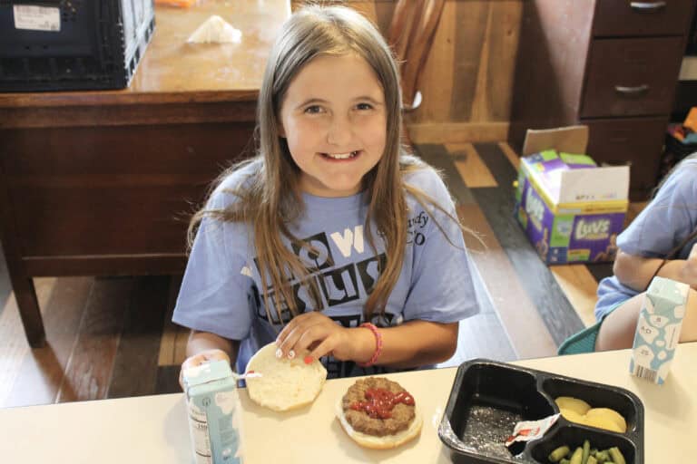 A young girl with long blonde hair smiles at the camera while preparing a hamburger at a table, with a milk carton, meal tray, and vegetables in front of her. She is wearing a light blue shirt.