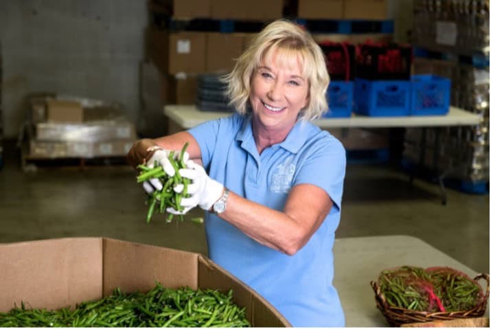 A woman wearing a blue polo shirt and white gloves smiles while sorting green beans in a warehouse, with crates and boxes in the background.