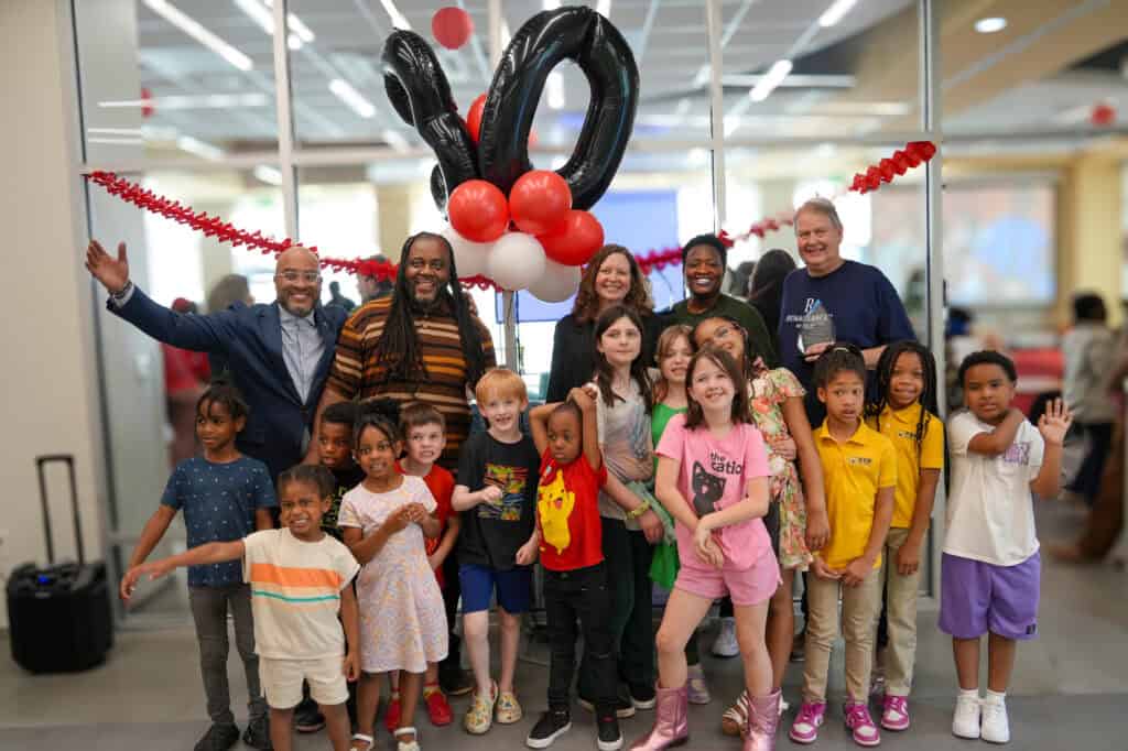 A group of adults and children smile and pose together indoors at a celebration. Black and red balloons shaped as 10 and festive decorations hang in the background.