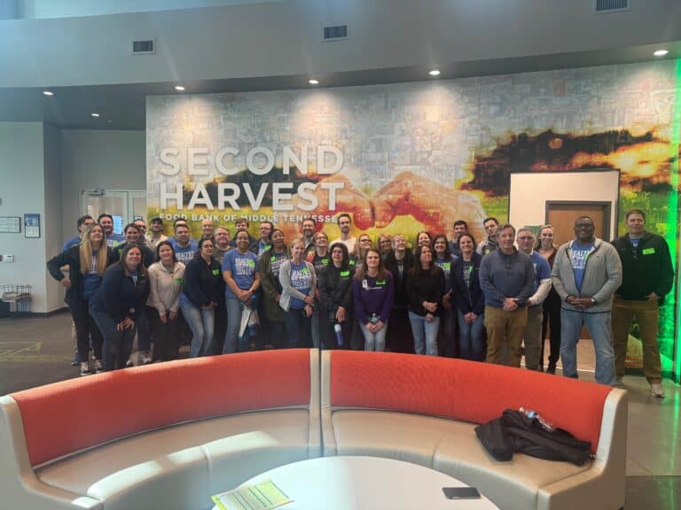 A large group of people pose and smile for a photo inside the Second Harvest Food Bank of Middle Tennessee, standing in front of a wall with the organization’s name and logo in a modern lobby with orange and gray furniture.