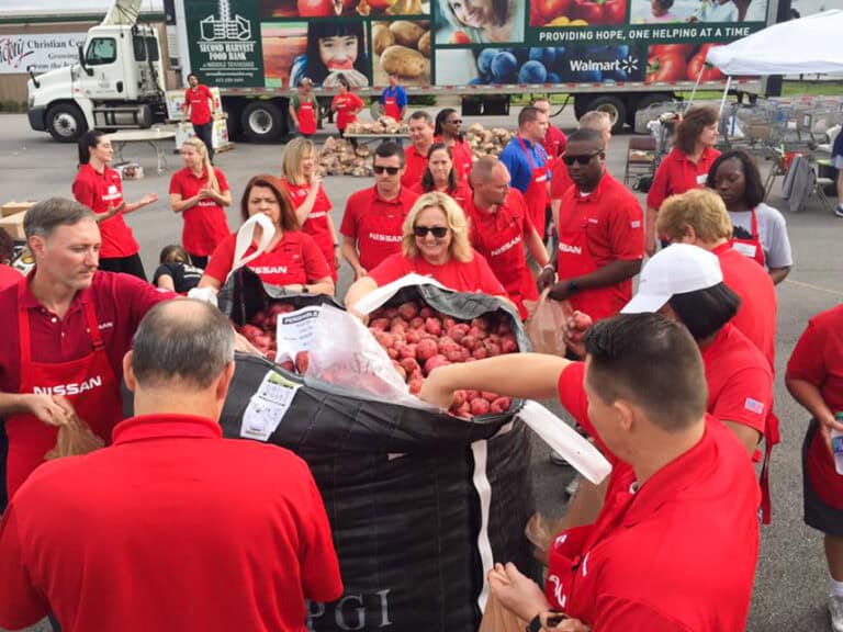 A group of people wearing red Nissan shirts gather around large bags of potatoes, packing them into smaller bags outdoors near a truck displaying produce and a Walmart logo.