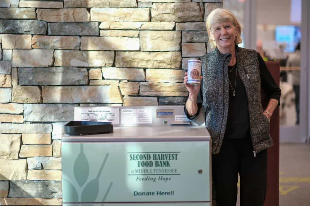 A woman stands smiling in front of a stone wall, holding a can of food beside a donation bin labeled Second Harvest Food Bank of Middle Tennessee. Donate Here! in a public indoor setting.